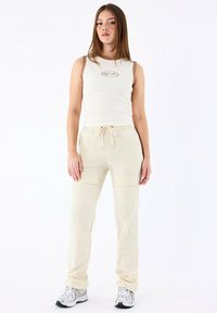 Pantaloni cargo beige con elastico in vita e cordoncino, tessuto strutturato e molteplici tasche; abbinati a un top aderente bianco con logo.