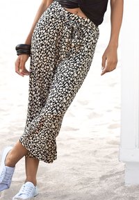 Kvinna som bär svarta och beigemönstrade cropped byxor, svart topp, vita sneakers och svarta armband, stående på en sandstrand.