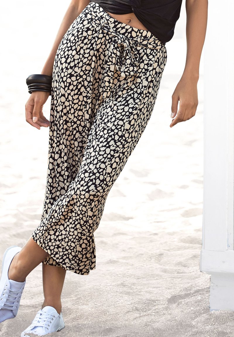 Kvinna som bär svarta och beigemönstrade cropped byxor, svart topp, vita sneakers och svarta armband, stående på en sandstrand.