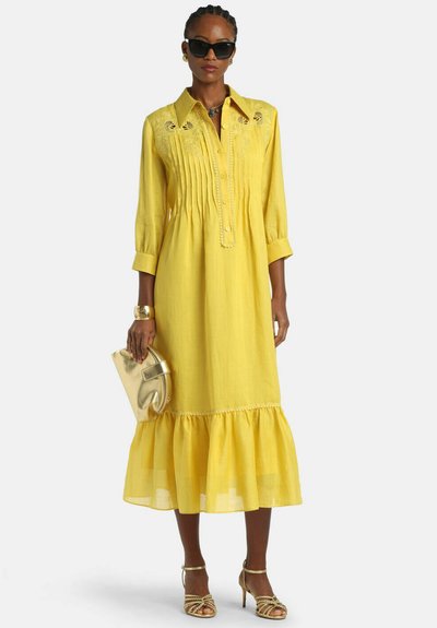 Žena v žltých midi šatách s plisovaním a výšivkami, drží zlaté psaníčko, má na nohách zlaté lodičky, čierne slnečné okuliare a má zapletené vlasy.