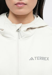 Veste à capuche beige clair avec une fermeture éclair intégrale et un logo "TERREX" gris. Texture lisse, design minimaliste, sans motifs ou accents remarquables.
