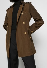 Person klädd i en brun dubbelknäppt trenchcoat med stora knappar över en svart polo och svarta byxor, med händerna i fickorna på rocken.