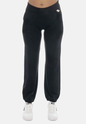 Personne portant un pantalon de jogging noir à taille élastique avec chevilles resserrées et des baskets blanches, debout devant un fond uni.