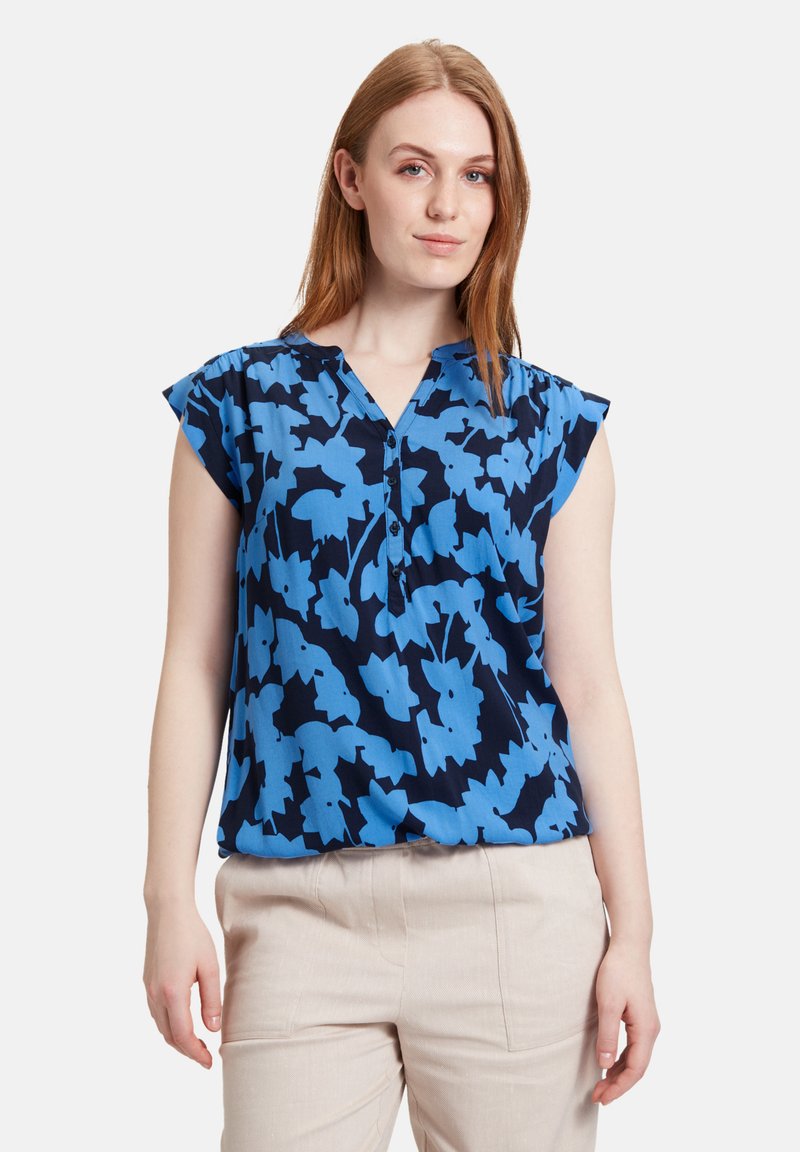 Blå bluse med blomsterprint, korte ærmer, V-udskæring og knapdetalje, lavet af let stof på en mørkeblå baggrund.