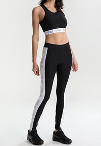 Kvinna som bär svart sportbh och svarta leggings med vita sidopaneler och FILA-logotyper, ihop med svarta träningsskor.