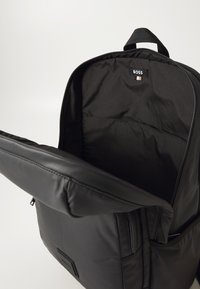 Sac à dos noir rembourré en matériau synthétique lisse, fermeture éclair et poche intérieure; il dispose d'une poignée sur le dessus et d'une étiquette de logo à l'intérieur.