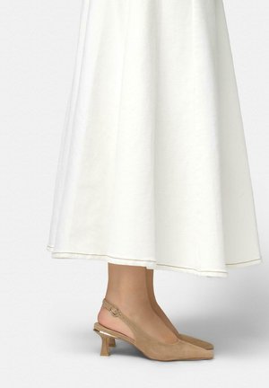 Piernas con zapatos de tacón bajo tipo kitten en ante beige con tiras traseras, combinados con una falda midi blanca sobre un fondo claro y liso.