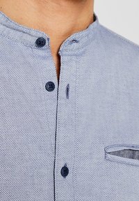 Camicia azzurro chiaro con colletto alla cinese, tessuto strutturato, due bottoni blu scuro e una piccola tasca sul petto.