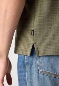 Pogled od blizu na osebo, ki nosi olivno zeleno majico z teksturo in oznako "BOSS" ter modre jeans hlače z vidnimi šivi.