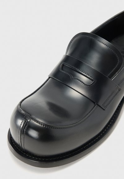 Crni kožni loafer s zaobljenim čelom, s prošivenim šavom i dekorativnom trakom preko gornjeg dijela. Glatka tekstura, minimalni akcenti.