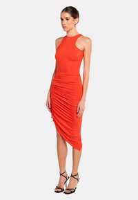 Rotes ärmelloses Kleid mit tailliertem Oberteil und asymmetrischem Saum, das seitliche Raffungselemente aufweist. Getragen mit schwarzen, dünnen High Heels.