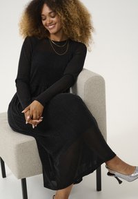 Black long-sleeve dress with a textured, sparkly fabric; accompanied by silver high-heeled shoes. Sitting on a light gray upholstered chair.