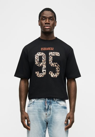 Άνδρας που φοράει μαύρο υπερμεγέθη μπλουζάκι με το λογότυπο "DSQUARED2" και μοτίβο λεοπάρδαλης με τον αριθμό "95" στο μπροστινό μέρος, συνδυασμένο με ανοιχτό μπλε ξεθωριασμένο τζιν.