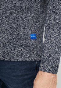 Granatowy sweter z dzianiny z teksturowanym wzorem i prążkowanym dołem. Na dolnej lewej stronie znajduje się mała niebieska naszywka z logo.