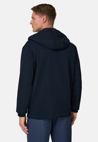 Mann steht mit dem Rücken zur Kamera, trägt einen dunkelblauen Hoodie mit offenem Kapuzen und dunkelblaue Hose vor einem schlichten, hellen Hintergrund.