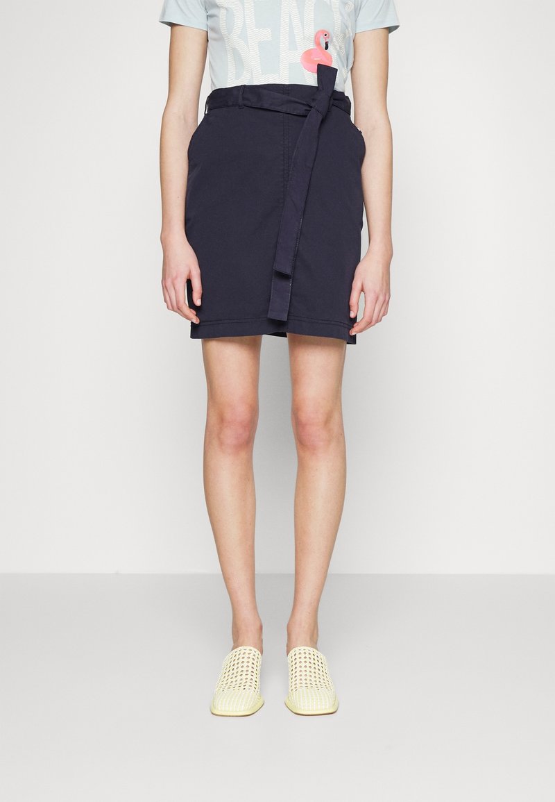 Personne portant une jupe bleu marine avec ceinture, un t-shirt bleu pâle « BEACH » avec un détail flamant rose, et des chaussures tissées crème à enfiler, debout devant un fond uni.