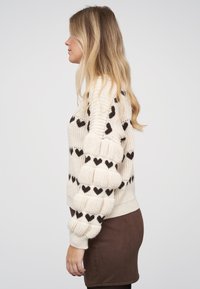 Blondýna v profilu, oblečená v krémovém svetru s černými srdíčkovými vzory a hnědé sukni na světlém jednobarevném pozadí.