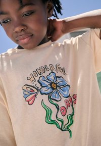 Lys beige t-shirt med farverigt grafisk design af en blå blomst, en sommerfugl og teksten "WONDERFUL DAYS" i rød.