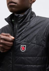 Asmuo užsisega juodą kimštinį džemperį su raudono lapės emblemą ant krūtinės priešais paprastą foną.