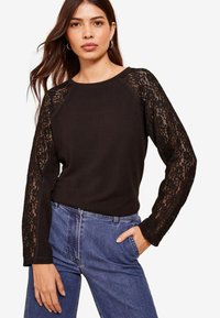 Maglietta nera a maniche lunghe con dettagli in pizzo sulle maniche, scollatura a girocollo e fit rilassato, abbinata a jeans in denim blu a vita alta.