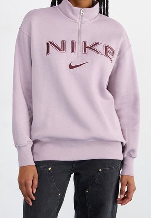 Person trägt ein hellviolettes Nike-Viertel-Reißverschluss-Sweatshirt mit dunkelrotem Logo, kombiniert mit schwarzen Jeans mit Metalldetailnieten.