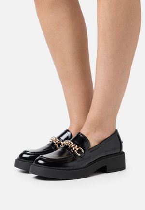 Mocassins en cuir verni noir avec une épaisse semelle noire compensée, dotés d'un accent en chaîne dorée à l'avant. Texture lisse, design moderne.