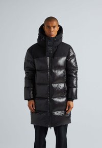 Cappotto puffo nero con finitura lucida, caratterizzato da un alto colletto, cappuccio e chiusura frontale con zip. Progettato con ampi pannelli orizzontali e dettagli con logo.