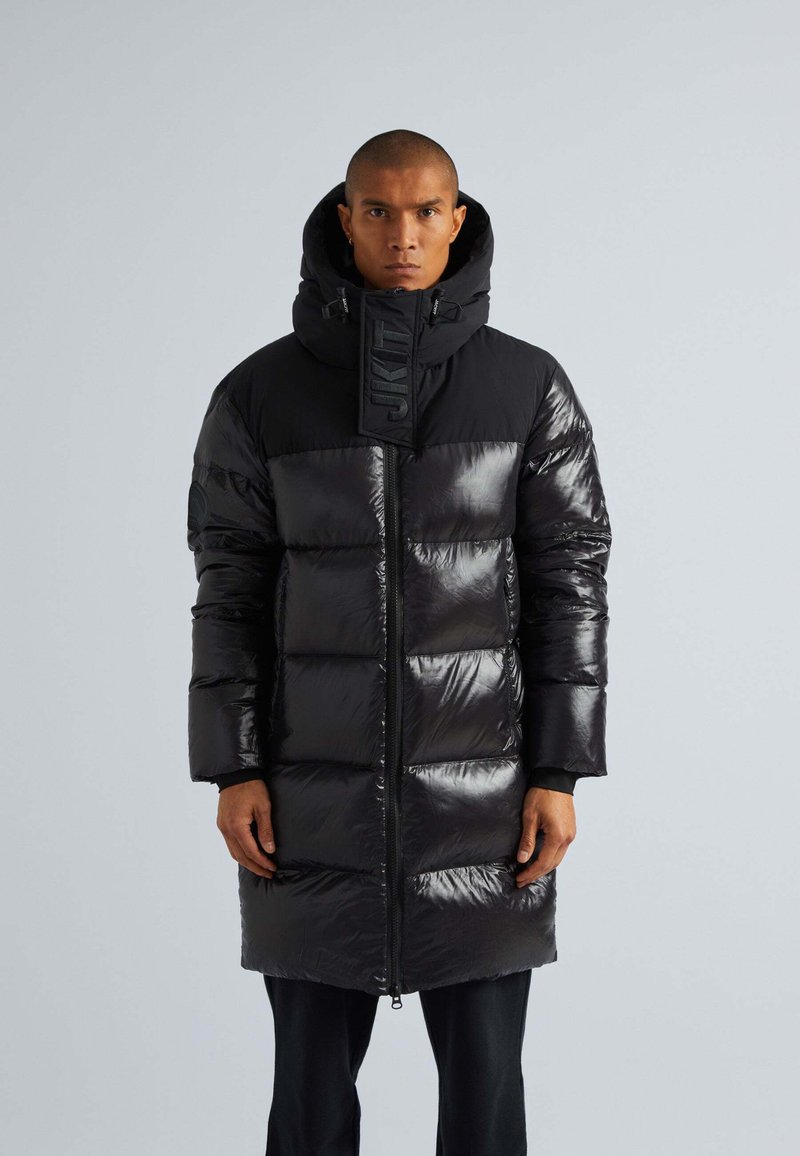 Cappotto puffo nero con finitura lucida, caratterizzato da un alto colletto, cappuccio e chiusura frontale con zip. Progettato con ampi pannelli orizzontali e dettagli con logo.