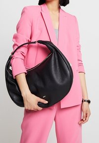 Kobieta w różowym garniturze trzymająca dużą czarną skórzaną torbę o zaokrąglonym kształcie i detalu zamka na jednolitym tle.