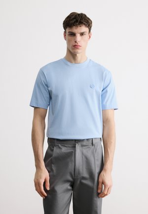 Νεαρός άνδρας φορώντας ανοιχτό γαλάζιο κοντομάνικο μπλουζάκι και γκρι παντελόνι, στέκεται μπροστά σε ένα απλό λευκό φόντο.