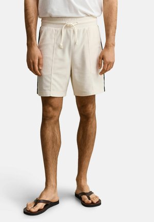 Mann trägt weiße Shorts mit Kordelzug, schwarzen seitlichen Streifen und braunen Flip-Flops, steht vor einem schlichten Hintergrund.