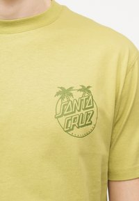 Ανοιχτό πράσινο μπλουζάκι με σκούρο πράσινο λογότυπο "Santa Cruz Skateboards" που απεικονίζει δύο φοίνικες στο στήθος.