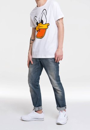 Persoon in een wit t-shirt met een grote cartoon-eendenface, blauwe opgerolde spijkerbroek, witte sneakers, staand tegen een eenvoudige witte achtergrond.