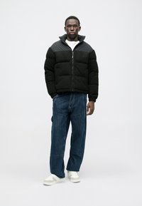 Casaco puffer preto com tecido texturado e ombros lisos, fecho com zip, combinado com jeans azuis de corte largo e ténis brancos.