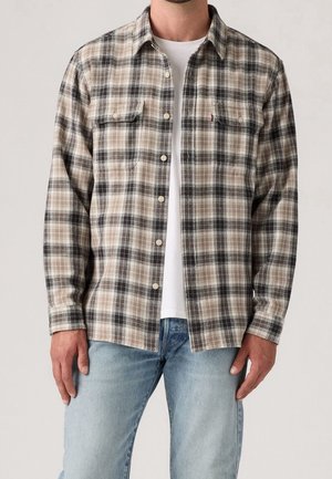 Homme portant une chemise à boutons à carreaux beige, noir et blanc par-dessus un t-shirt blanc avec un jean bleu clair, debout devant un fond uni.