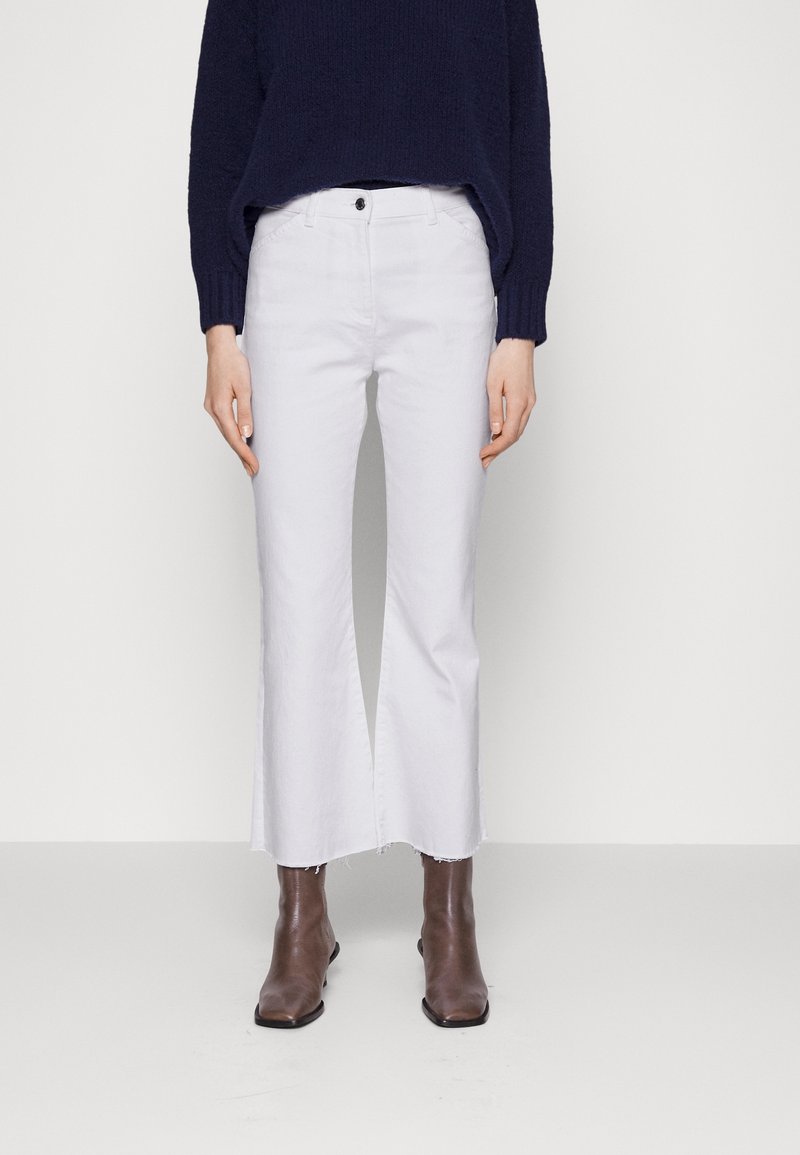 Vrouw draagt witte, hoog getailleerde, wijd uitlopende cropped broek met rafelige zoom, een donkerblauwe trui en bruine enkellaarzen.