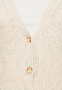 Μπεζ ζακέτα από ριμπ πλεκτό με δύο ανοιχτό καφέ κουμπιά πάνω από λευκό ριμπ πουκάμισο.