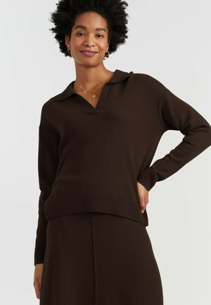 Donna con capelli corti e ricci, che indossa un maglione marrone scuro a maniche lunghe con colletto e gonna abbinata, sorridente con una mano sui fianchi.