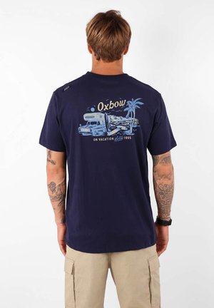 T-shirt en coton bleu marine avec un graphisme d'un campeur et de palmiers, portant l'inscription "Oxbow EN VACANCES depuis 1985" imprimée en bleu clair.