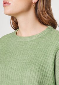 Gerippter grüner Pullover mit rundem Ausschnitt, gefertigt aus einem weich strukturierten Stoff. Goldene Creolen akzentuieren den Look.