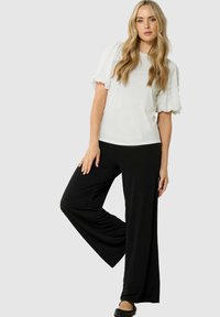 Donna con lunghi capelli biondi che indossa un top bianco con maniche a sbuffo, pantaloni neri a gamba larga e ballerine nere, in posa contro uno sfondo grigio chiaro.