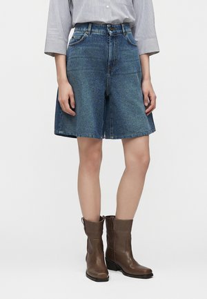 Person iført løse denimshorts til knæene, en stribet skjorte med opkrogede ærmer og brune læderstøvler til midt på læggen.
