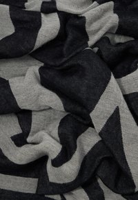 Strikket skjerf i svart og grått, med et geometrisk mønster og diagonale striper. Myk tekstur, lettvektig, og henger løst.