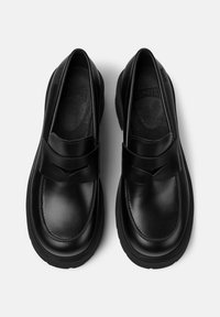 Un par de mocasines de cuero negro con suelas gruesas y una correa ancha en la parte superior, mostrados desde arriba sobre un fondo blanco.