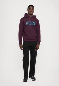 Μωβ φούτερ με το λογότυπο "VERSACE JEANS COUTURE" σε τυρκουάζ, συνδυασμένο με μαύρα tapered παντελόνια και μαύρα αθλητικά παπούτσια. Απαλή υφή, άνετη εφαρμογή.