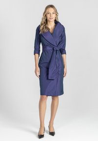 Granatowa sukienka zapinana na wstążkę do kolan, z szerokim kołnierzem, rękawami o długości trzech kwart i detalem w postaci związanego w pasie. Gładka tekstura i dopasowany fason.