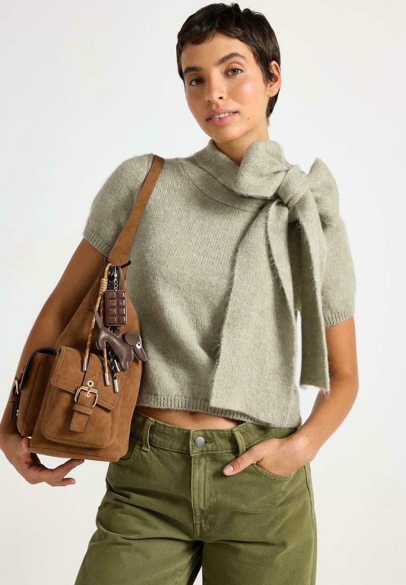 Ljusgrön, kortärmad tröja med en rosettdetalj vid halsringningen. Kombinerad med olivgröna byxor och en brun mockaväska med metalldetaljer.