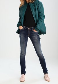 Tyrkysová bomber bunda přes černý top, doplněná o tmavě modré úzké džíny a světle růžové boty. Bunda má hladkou, lesklou texturu.