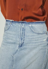 Jupe en denim bleu clair avec une taille haute, un ourlet effiloché et en détresse, et deux poches avant. Assortie à une chemise à boutons orange-brun.