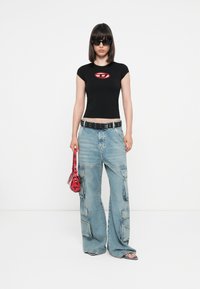 Black fitted T-shirt with red logo, baggy light blue denim cargo pants, black belt, and small transparent red handbag. Black sunglasses.